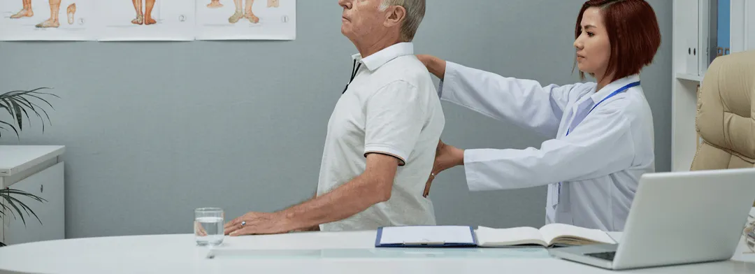 A healthcare professional wearing a white coat examining the back of an older man seated at a desk in a medical office.