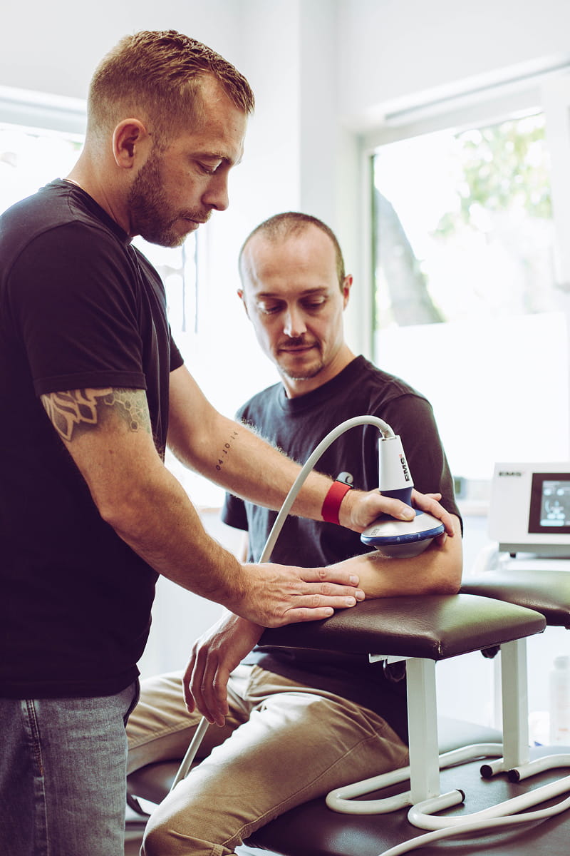 A male therapist uses a handheld ultrasound therapy device on a seated man's forearm in a clinical setting, with sunlight coming through a window in the background.