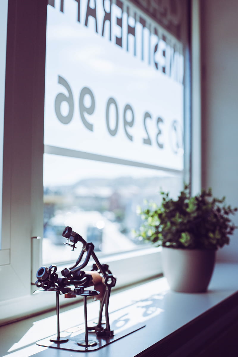 Metal sculptural figurine of a person working on a piece of machinery, placed on a windowsill with sunlight casting shadows, next to a potted green plant.