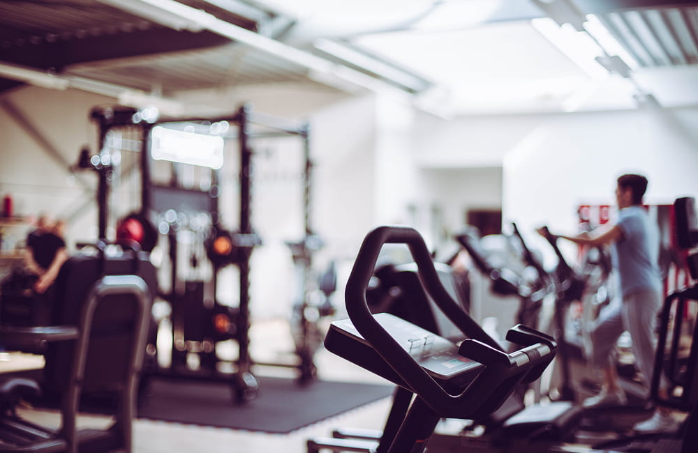 Modern gym with various exercise machines including stationary bikes and weightlifting equipment with a person exercising in the background.