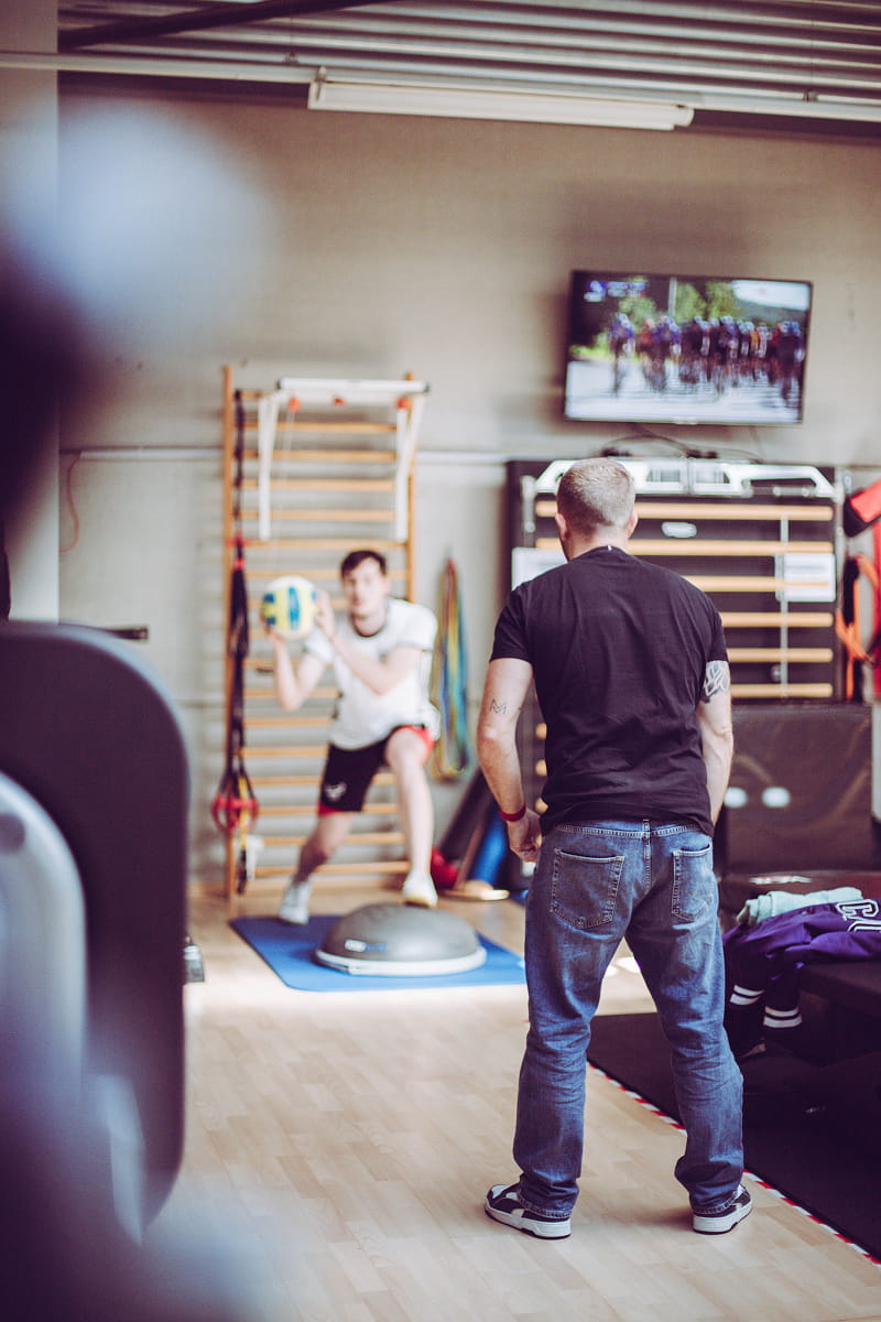 Man in a black shirt and jeans supervises another man in athletic wear holding a volleyball while doing a lunge exercise on a blue mat with a balance trainer in a gym room with wooden floor and a TV showing a cycling race on the wall.