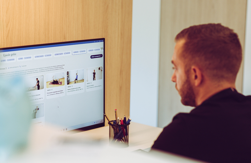 Man with short brown hair and beard looking at a computer screen displaying exercise tutorial thumbnails and French text in a wooden-paneled room.