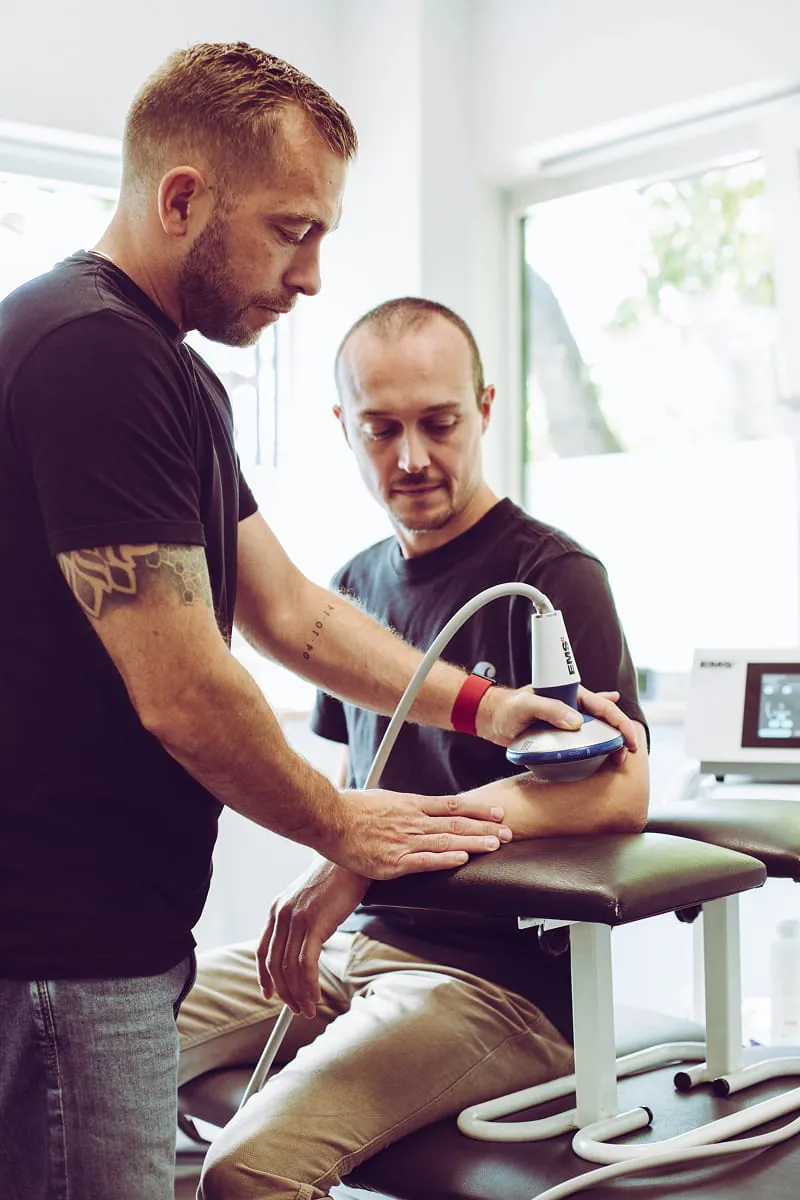 A male therapist uses a handheld ultrasound therapy device on a seated man's forearm in a clinical setting, with sunlight coming through a window in the background.
