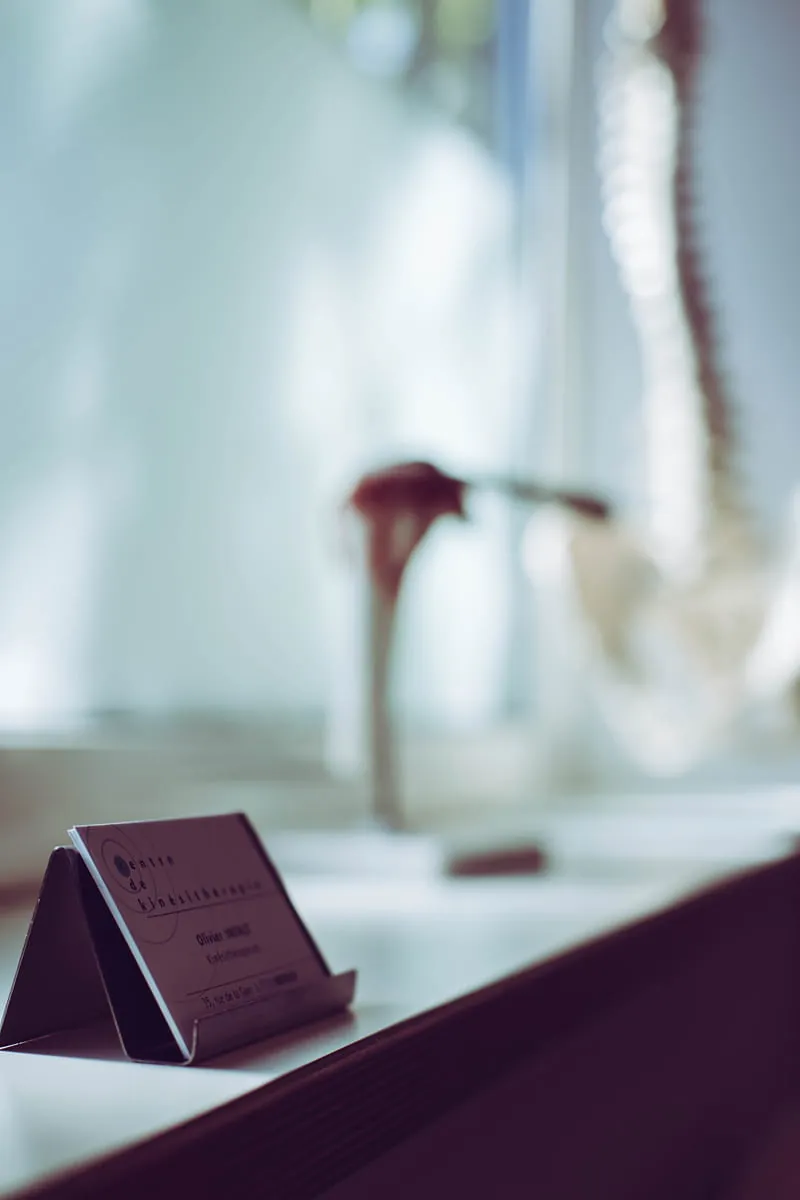 Close-up of a black business card holder with a card for a center of kinesitherapy, placed on a white surface next to a blurred anatomical skeleton model in the background.