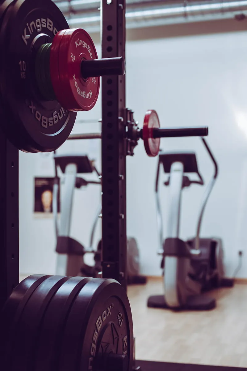 Close-up of a gym rack loaded with various Kingsbox weight plates in red, green, and black, with blurred exercise machines in the background.