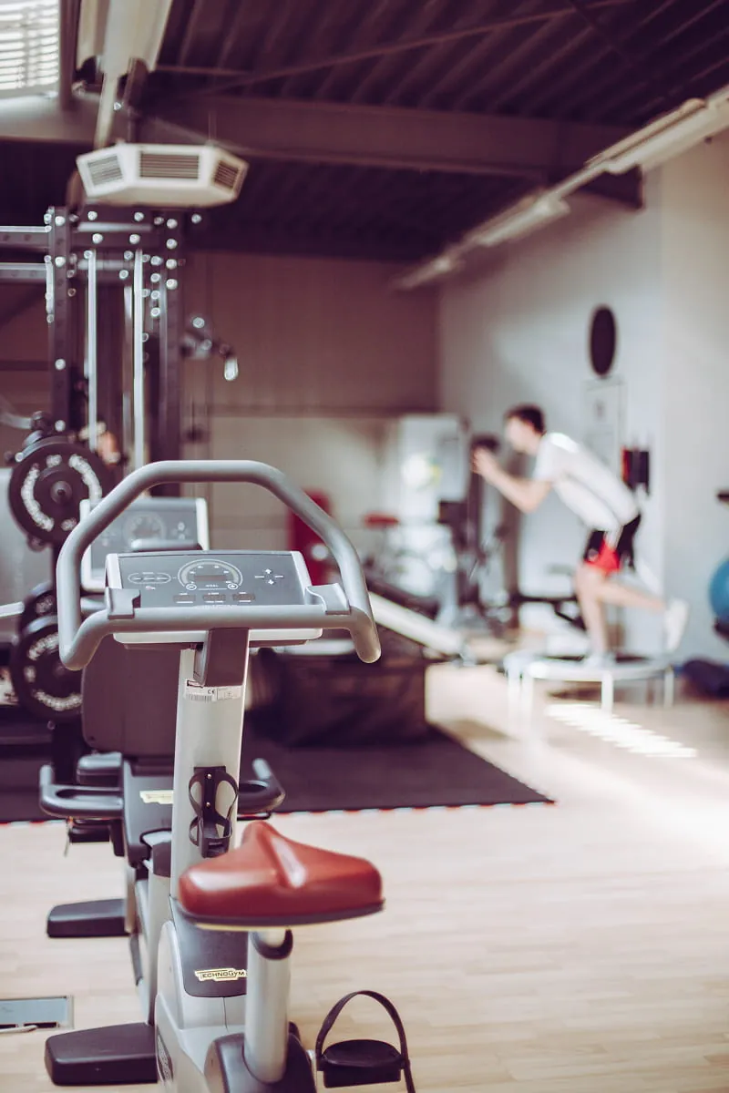 Indoor gym scene focusing on a stationary exercise bike with a digital display and brown seat in the foreground, while a person in white shirt and black shorts exercises on a trampoline in the blurred background.