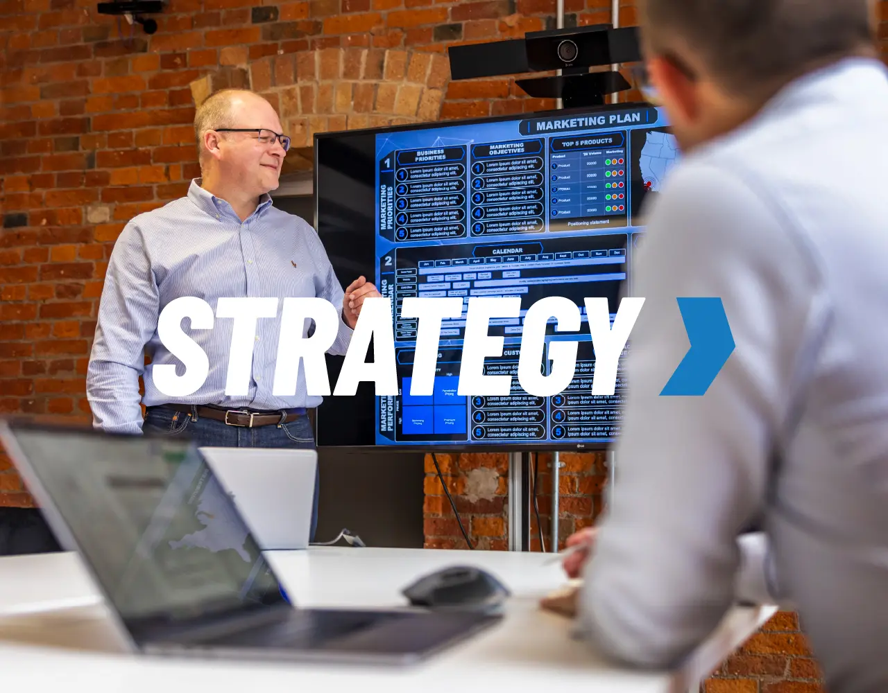 A man in a blue shirt is standing in front of a laptop screen with the word strategy on it.