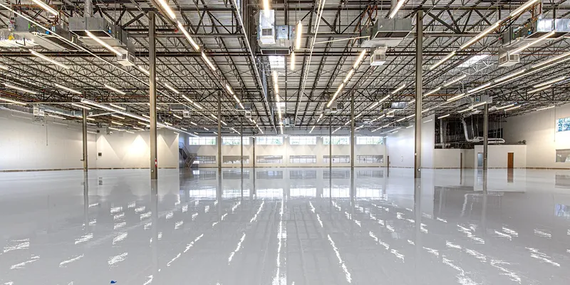 Große, leere Industriehalle mit glänzendem, reflektierendem Boden und sichtbarer Deckenstruktur.