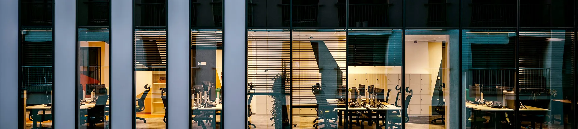 Blick durch Glasfenster auf ein menschenleeres Büro mit Schreibtischen und Computerarbeitsplätzen.