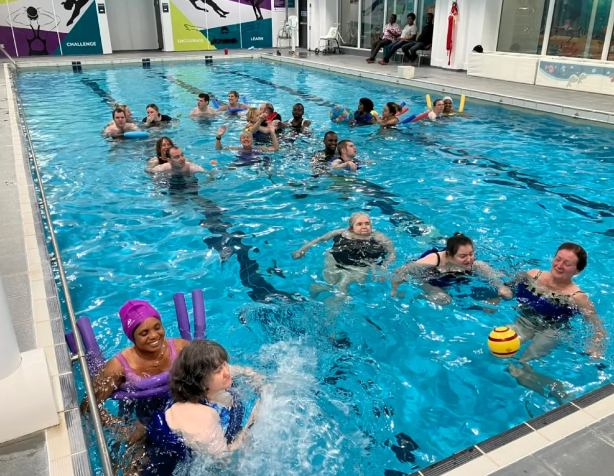 Group of adults enjoying a swimming pool session with pool noodles and a ball, indoors.