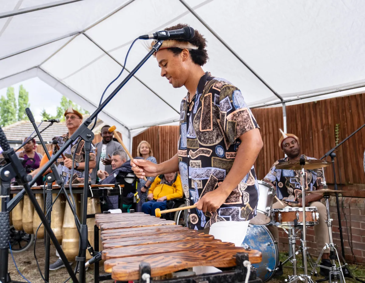 Musicians playing marimba and drums under a tent at an outdoor event with seated audience.