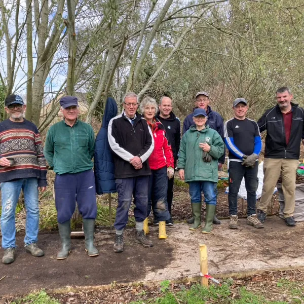 Group of nine adults standing outdoors on a dirt path surrounded by trees and bushes.