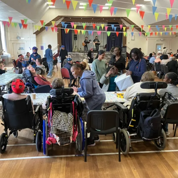 People in wheelchairs and caregivers socialise in a decorated hall with musicians performing on stage.