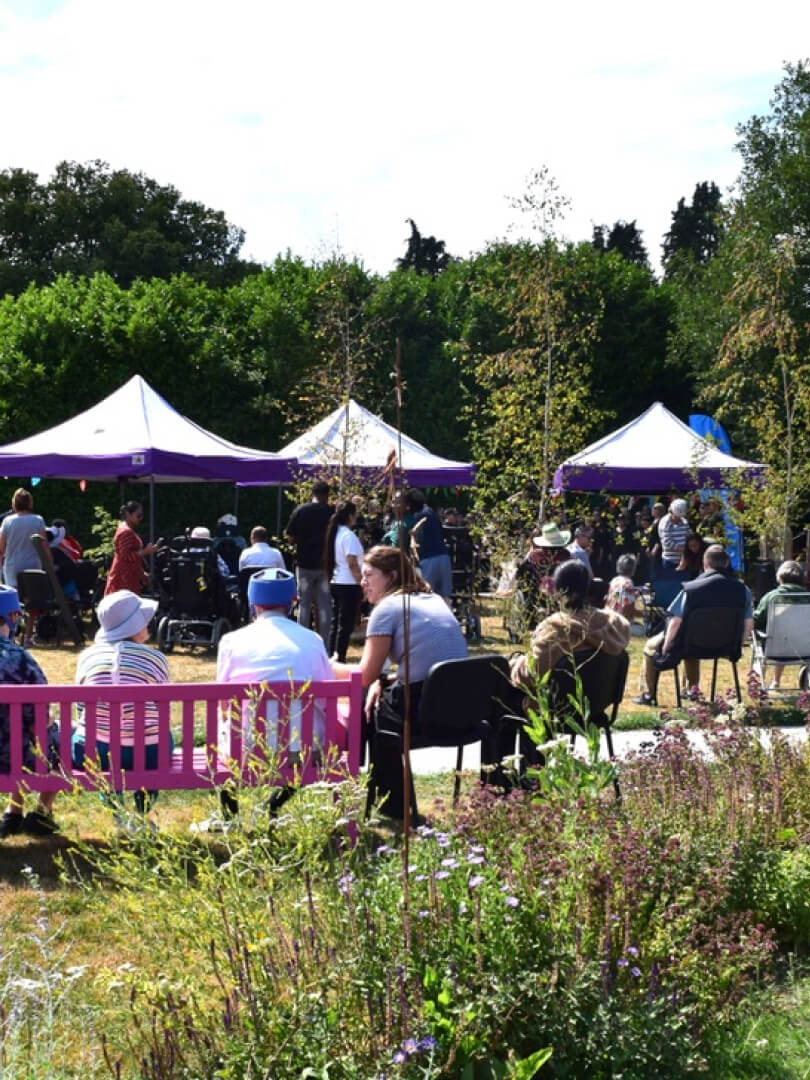 People gathered outdoors sitting under white tents and on chairs in a garden with green trees and flowers.