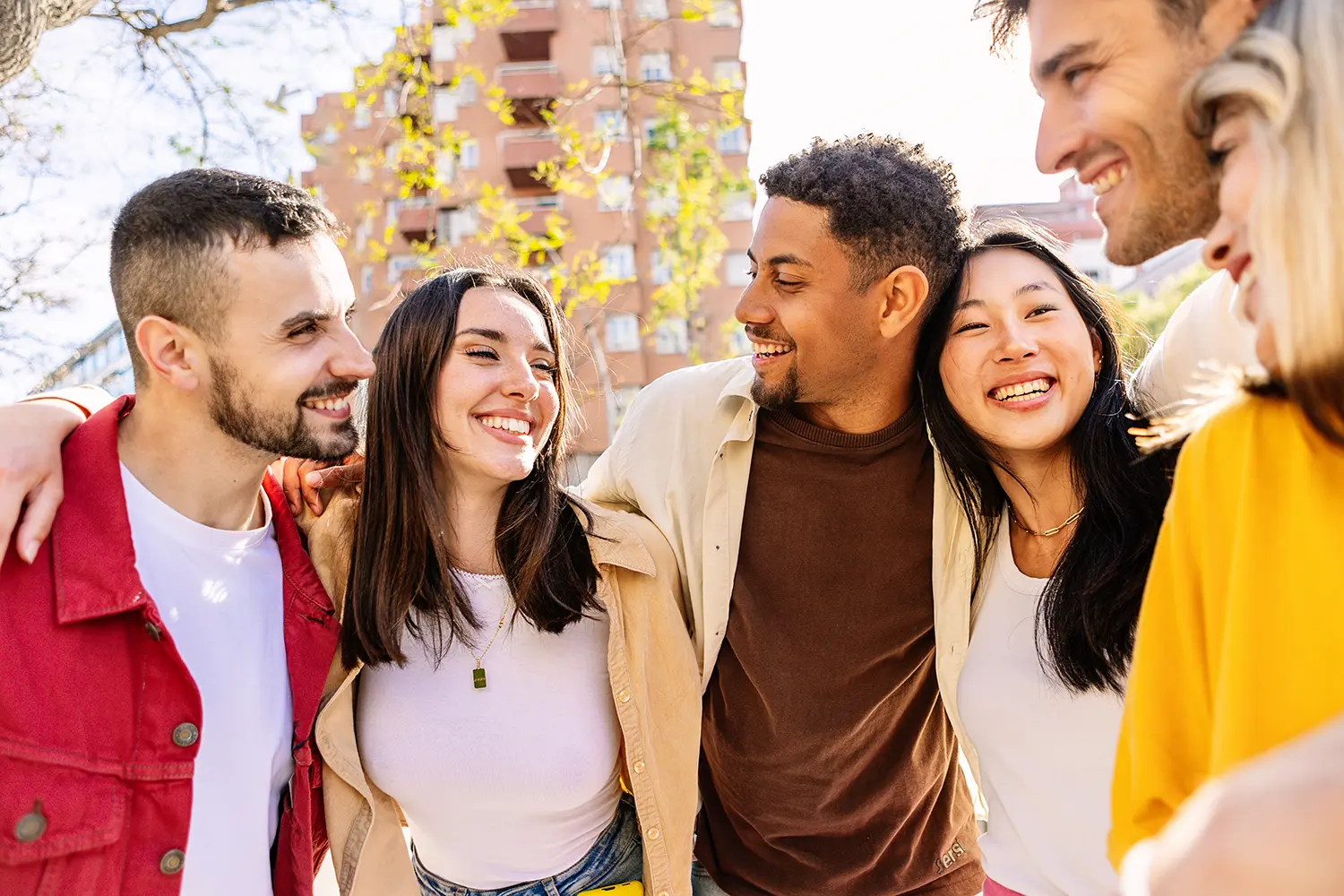 Group of five diverse young adults smiling and embracing outdoors in a sunny urban setting.