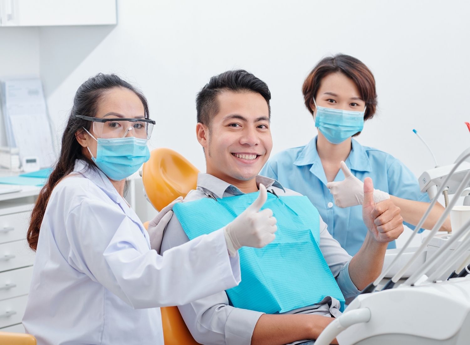 Smiling dental patient and two masked dental professionals giving thumbs-up in a clinic.