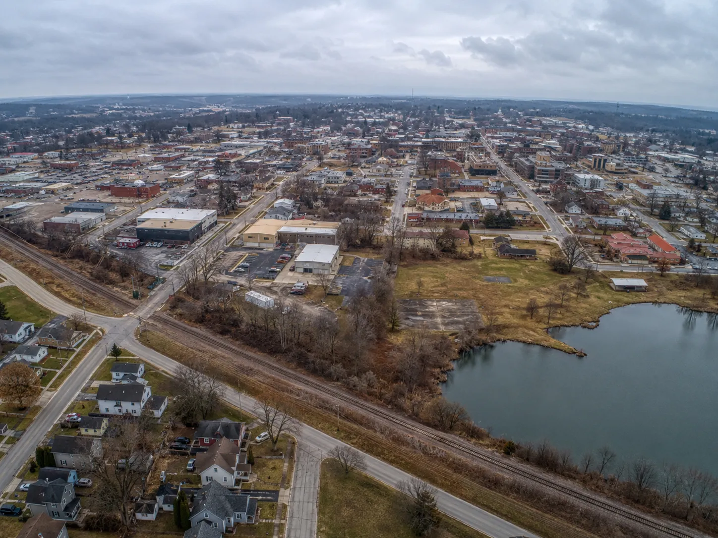 Aerial view of a town with houses, buildings, roads, a railroad, and a pond under a cloudy sky.