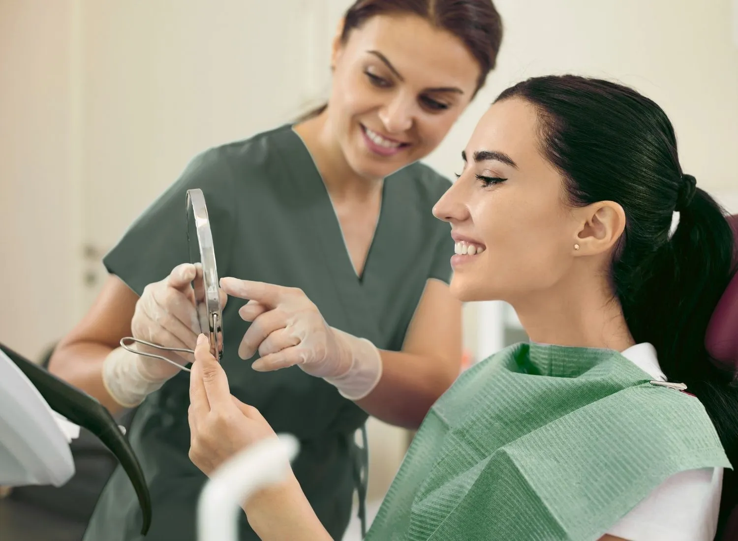 Smiling dental patient holding a mirror while a dental hygienist explains a procedure.