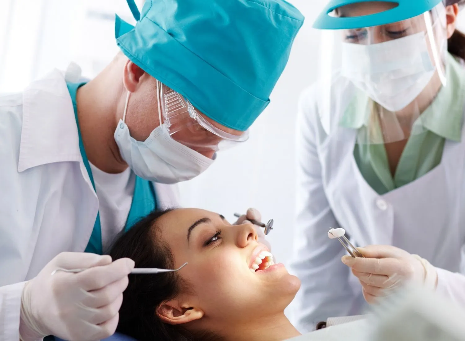 Dentist examining smiling female patient's teeth with dental assistant holding a cotton roll with tweezers.