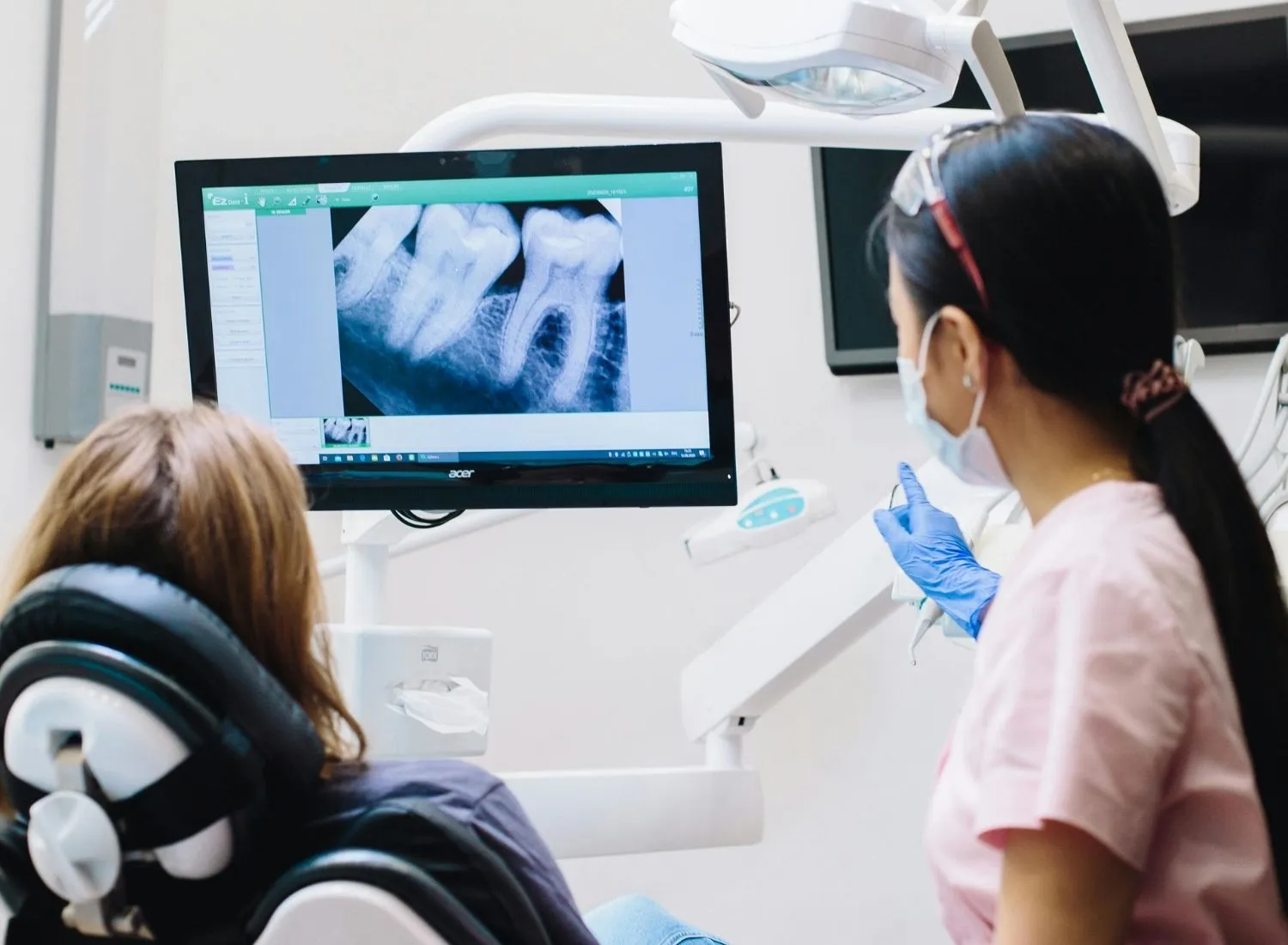 Dentist wearing mask and gloves showing a dental X-ray on screen to a patient in a chair.