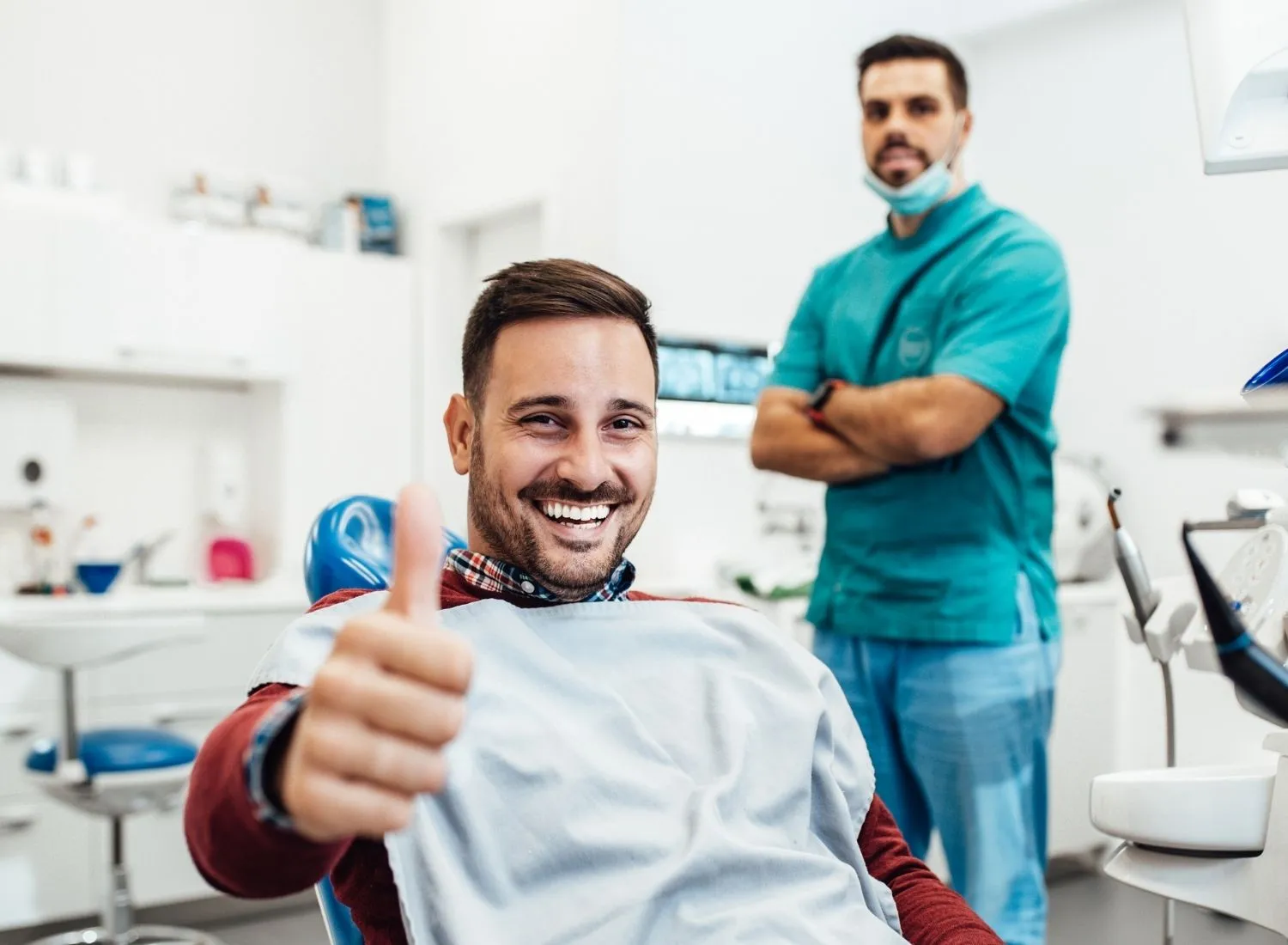 Smiling man in dental chair giving thumbs up with dentist standing in background arms crossed.