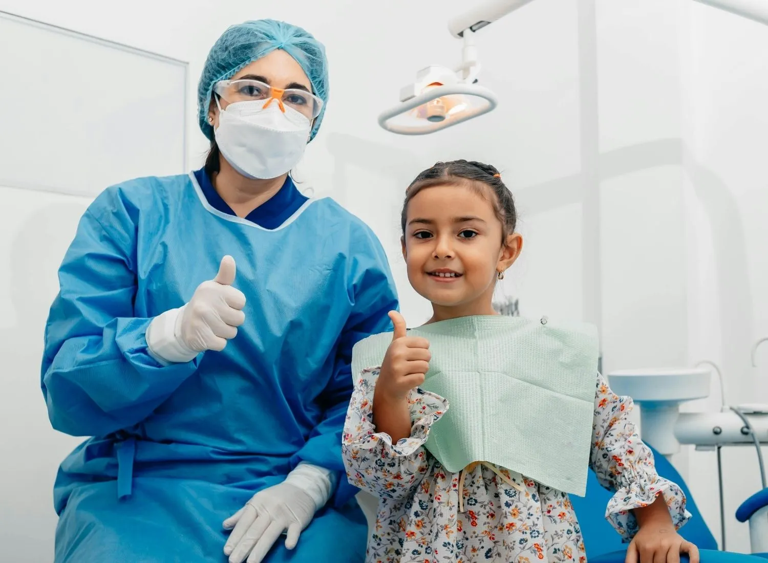 Dentist in protective gear and young girl patient both giving thumbs up in dental office.