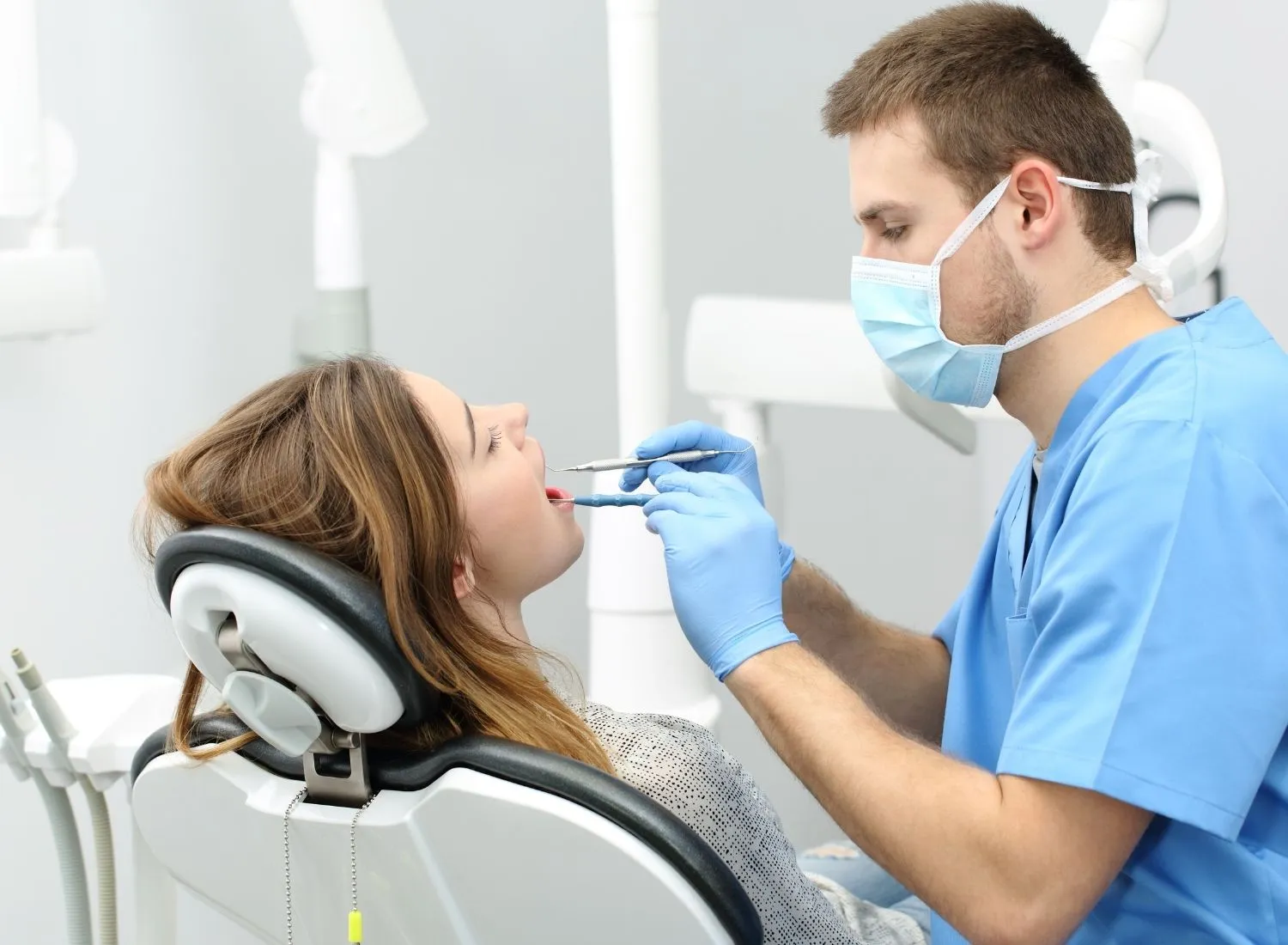Dentist wearing mask and gloves examining a female patient's mouth in a dental chair.