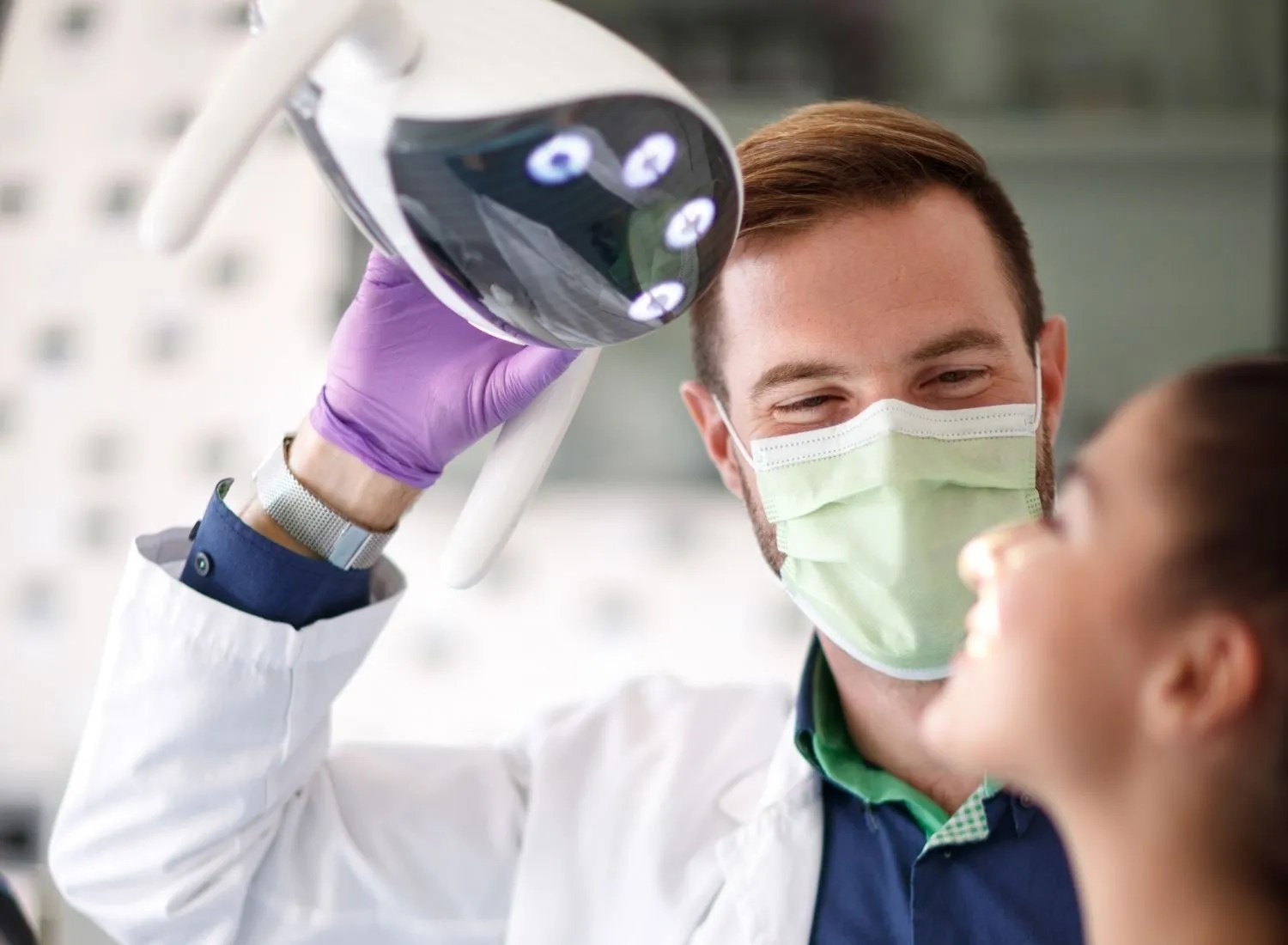 Dentist wearing a face mask and purple gloves adjusting a dental light over a patient's chair.