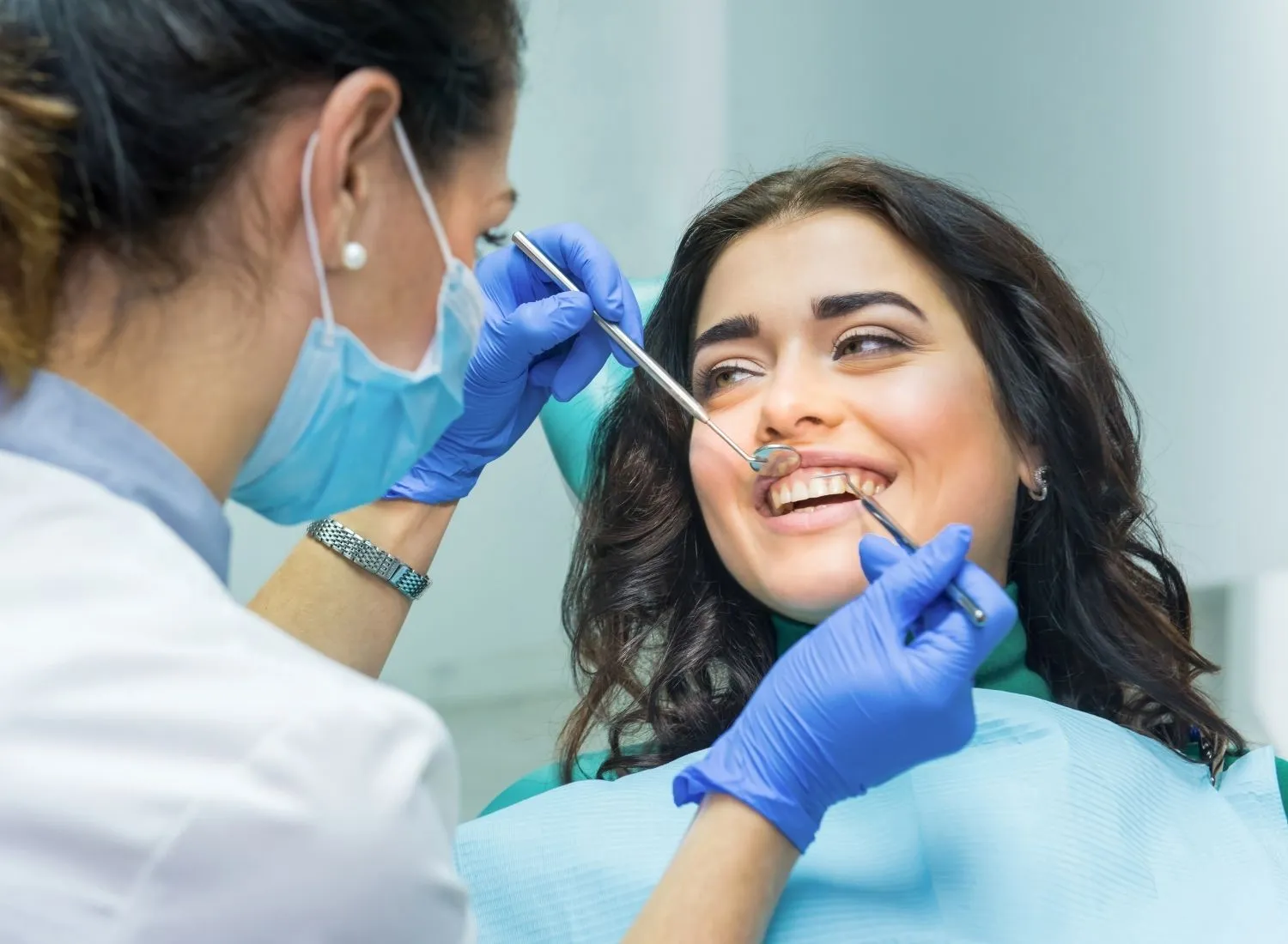 Dentist wearing mask and gloves examining a smiling patient's teeth with dental tools.