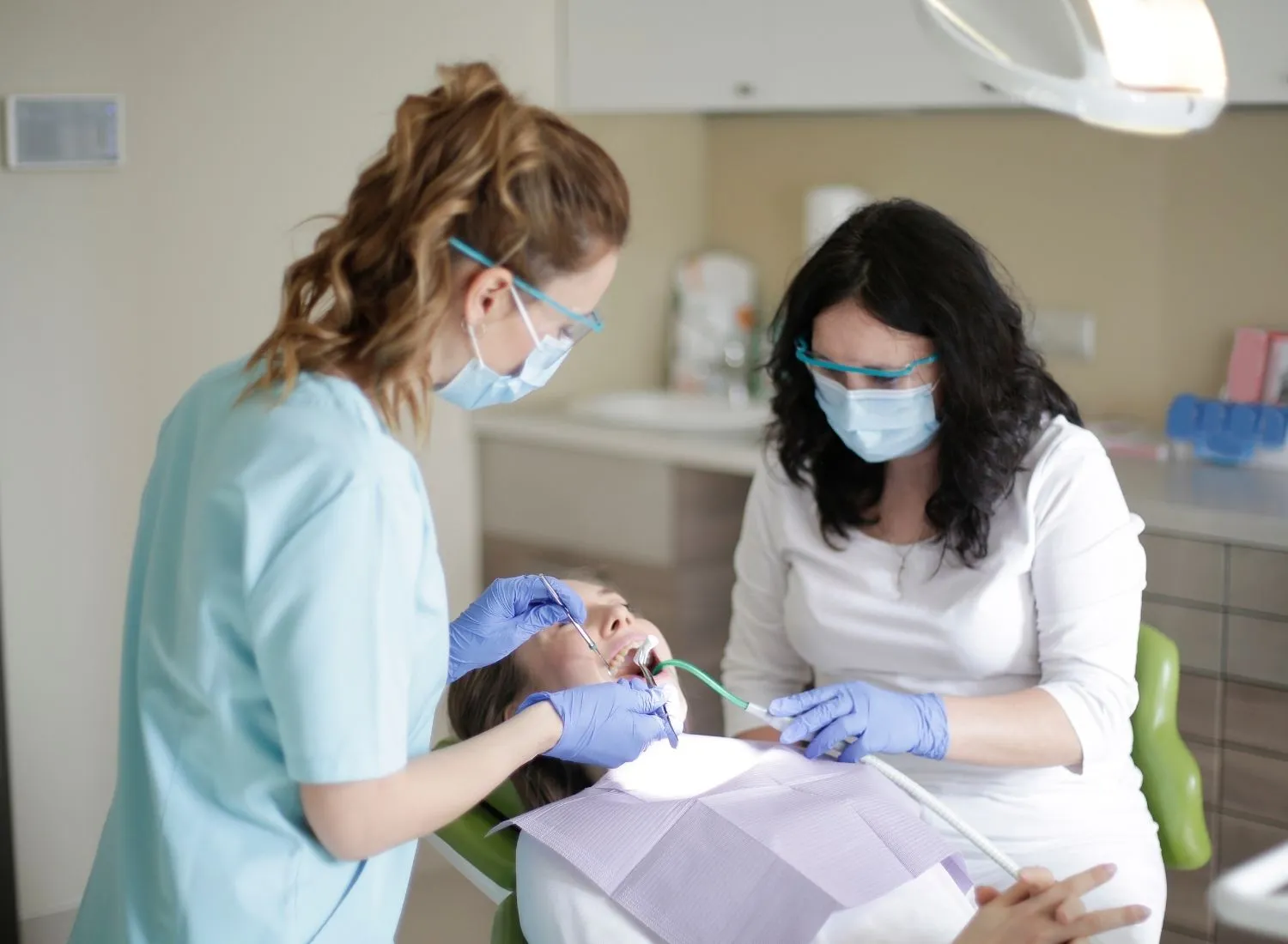 Two dental professionals wearing masks and gloves treating a patient reclining in a dental chair.