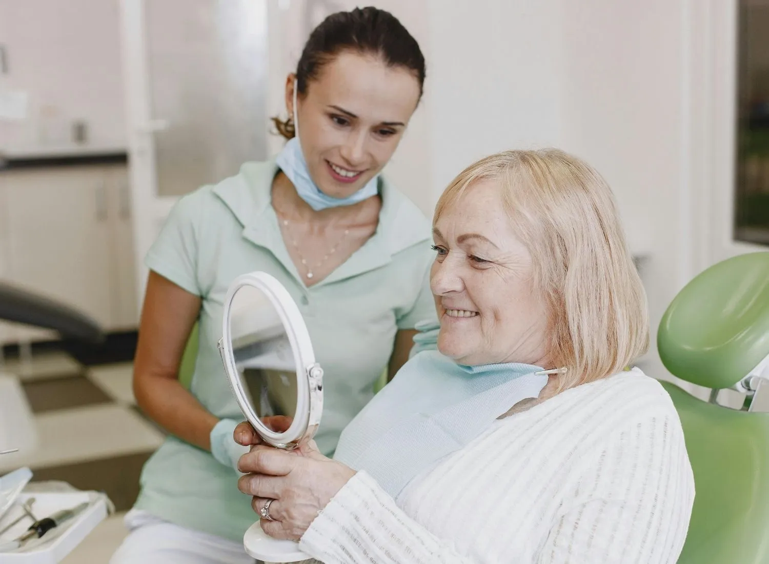 Smiling elderly woman in dental chair holding mirror with a dentist standing behind her.