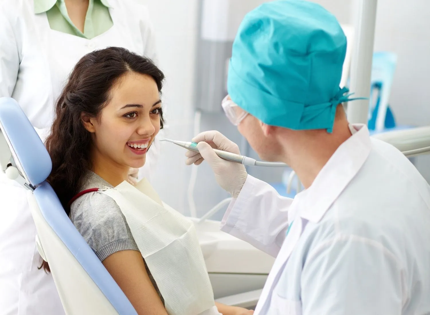 Dentist in blue surgical cap examining smiling young woman in dental chair with dental tool.