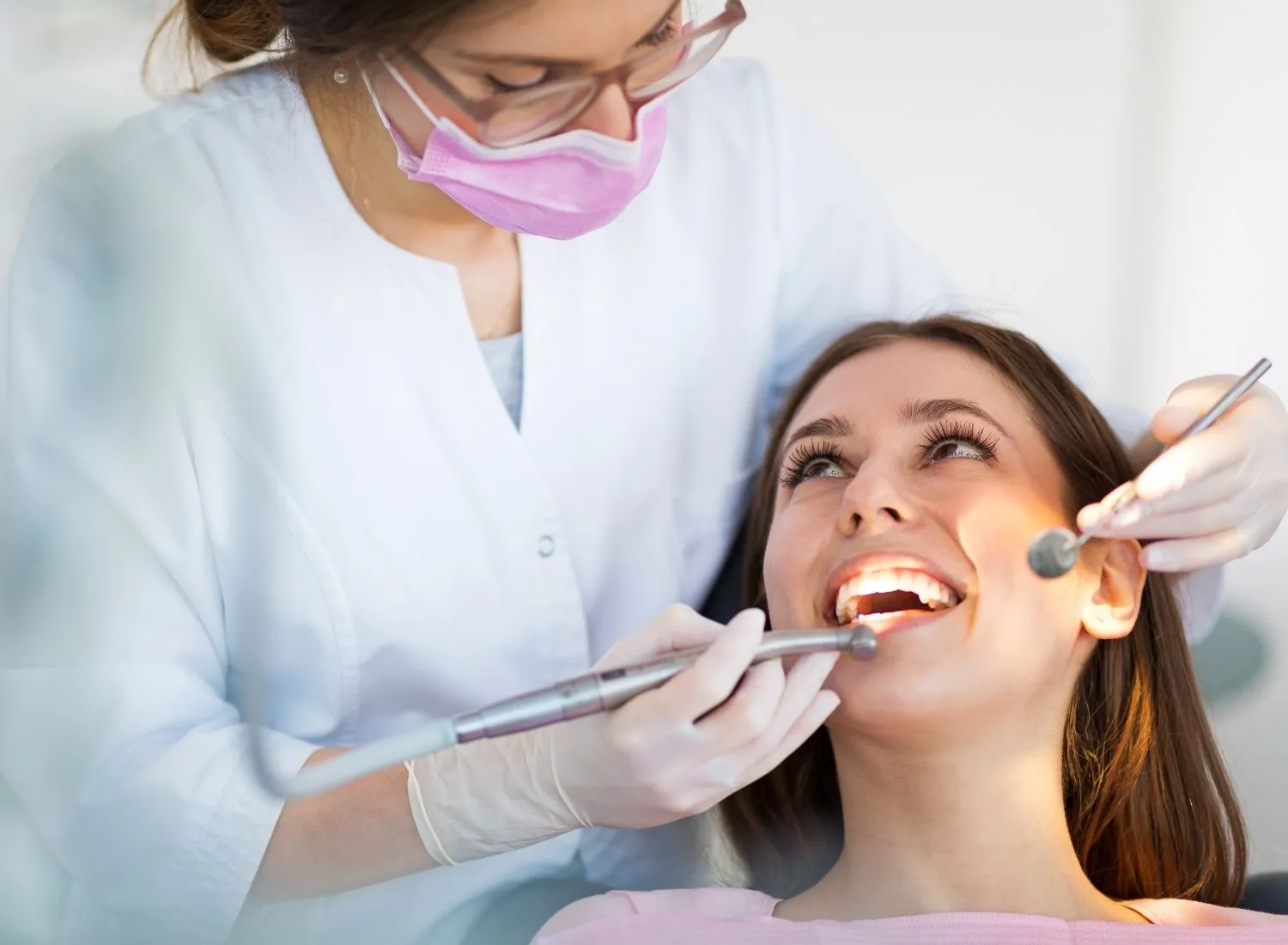 Dentist wearing mask examining smiling patient's teeth with dental tools in a bright clinic.