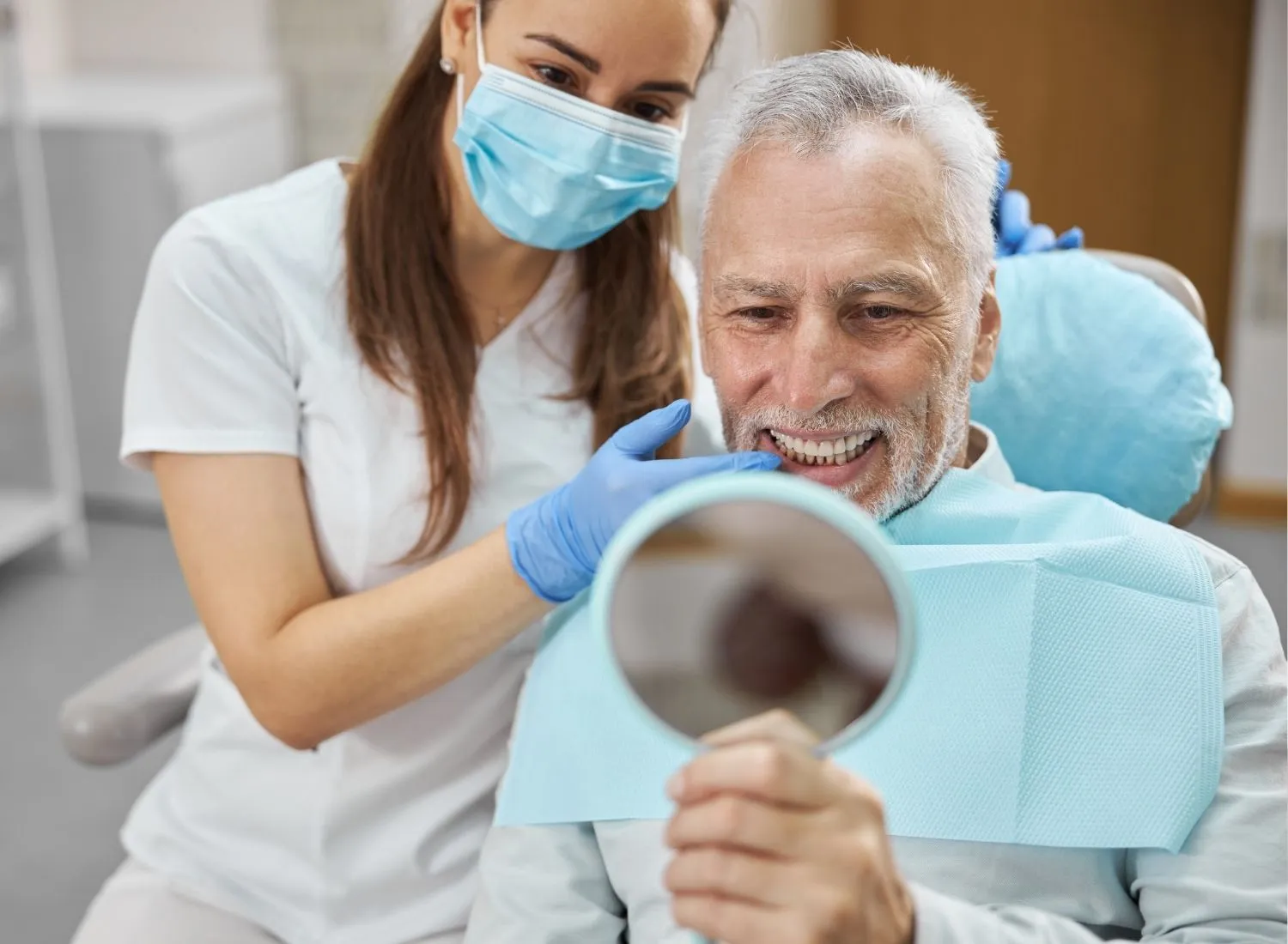 Male patient smiling and looking in a handheld mirror with a female dentist wearing a mask and gloves.