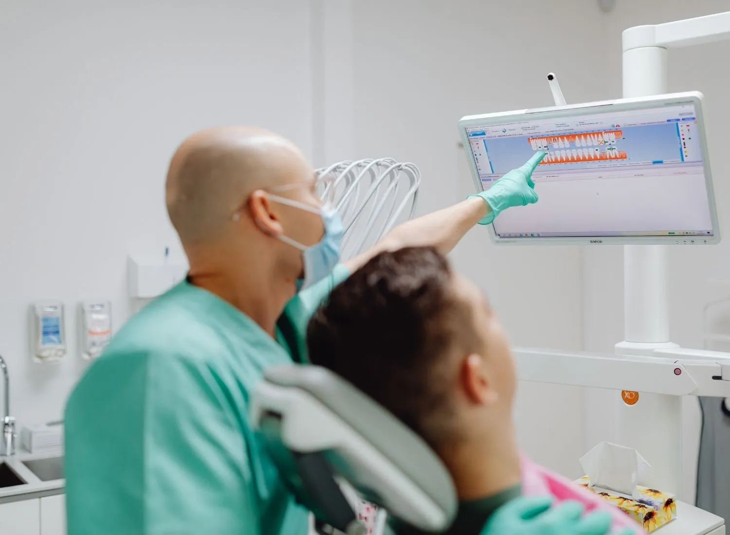 Dentist wearing mask and gloves points at dental X-ray on screen while patient sits in chair.