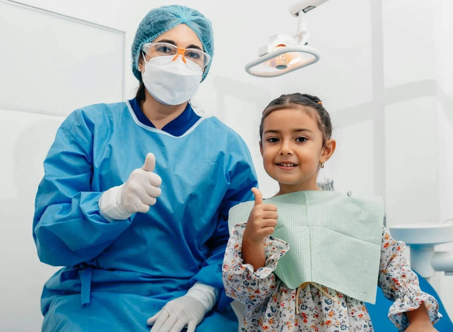 Dentist in protective gear and a young girl patient both showing thumbs up in a dental clinic.