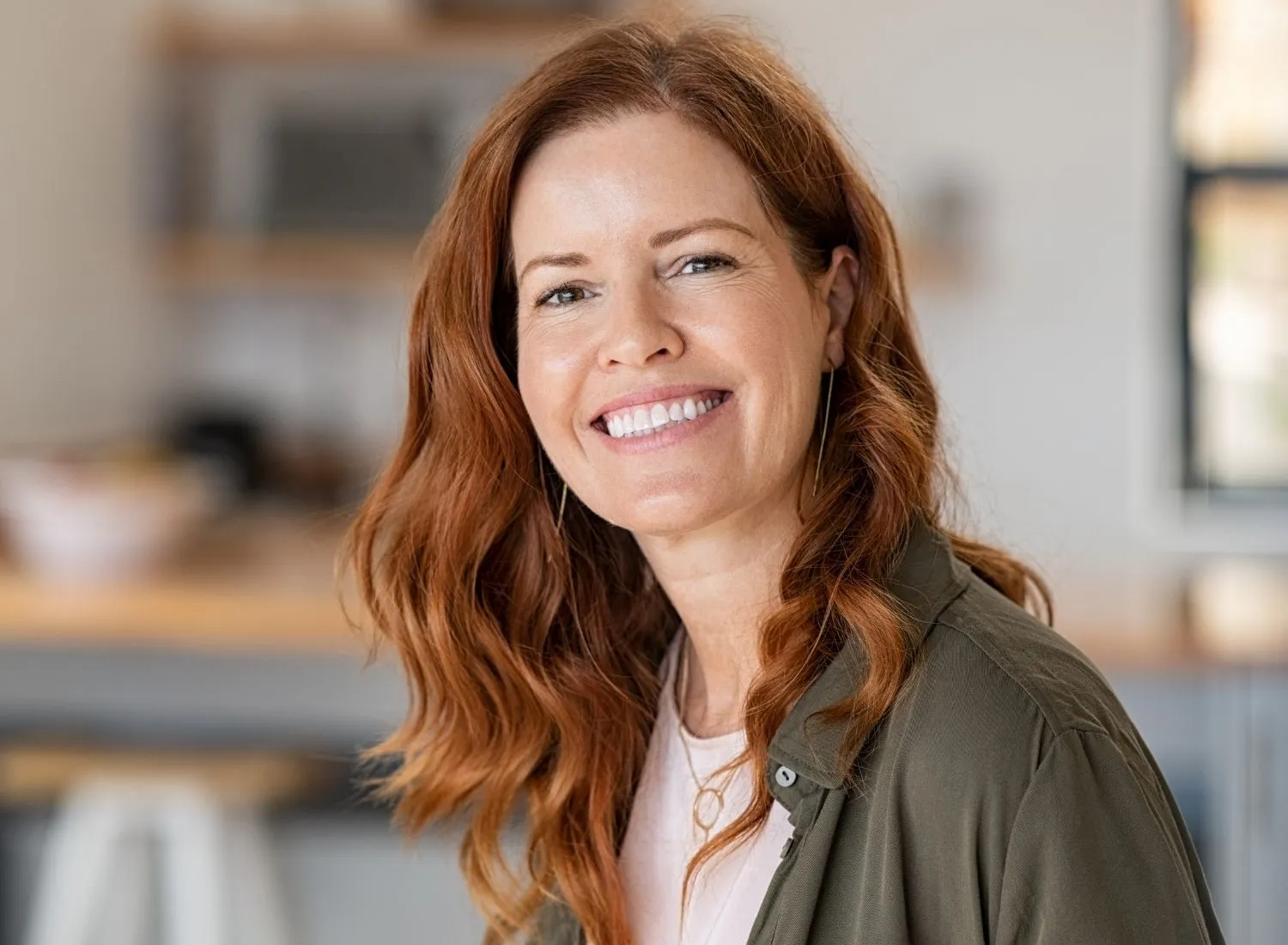 Smiling red-haired woman with wavy hair wearing a green shirt and gold necklace indoors.