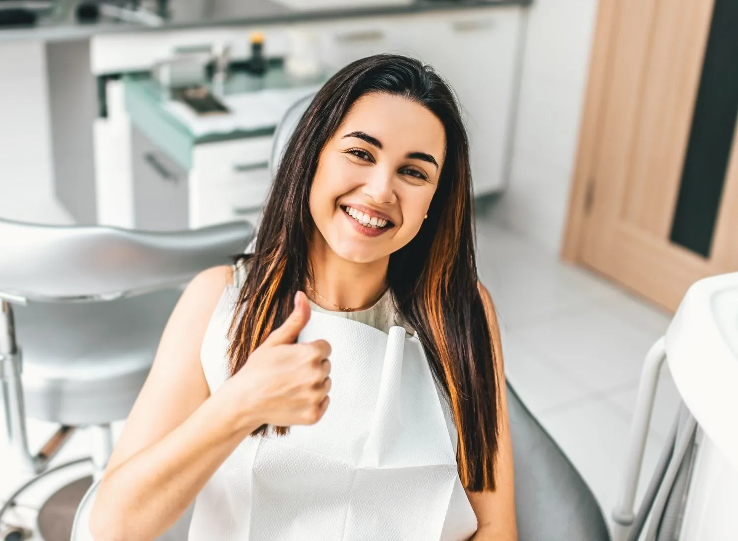 Smiling woman in dental chair giving thumbs up, wearing dental bib in a bright clinic.