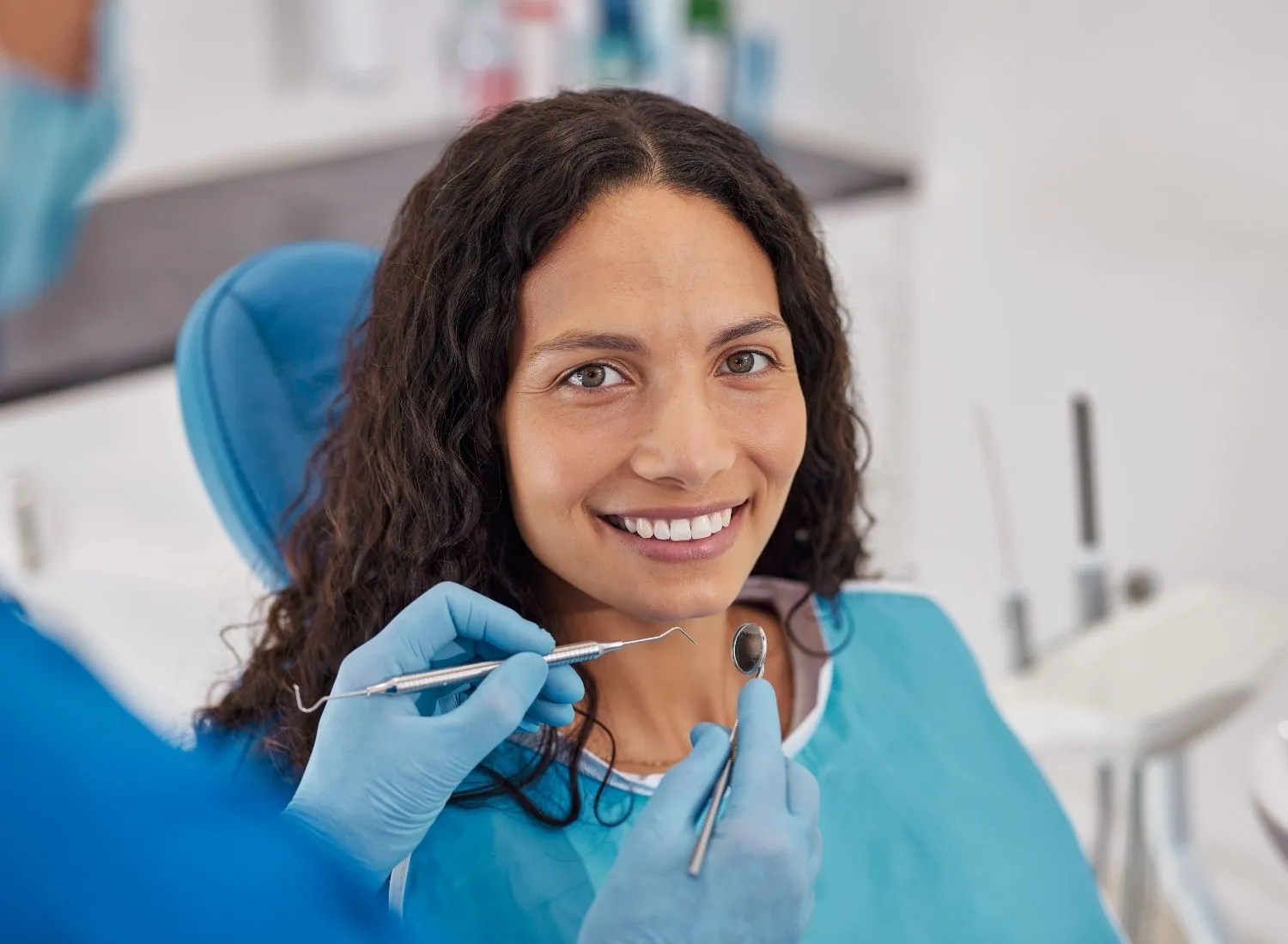 Smiling woman in dental chair being examined with dental mirror and pick by a gloved dentist.