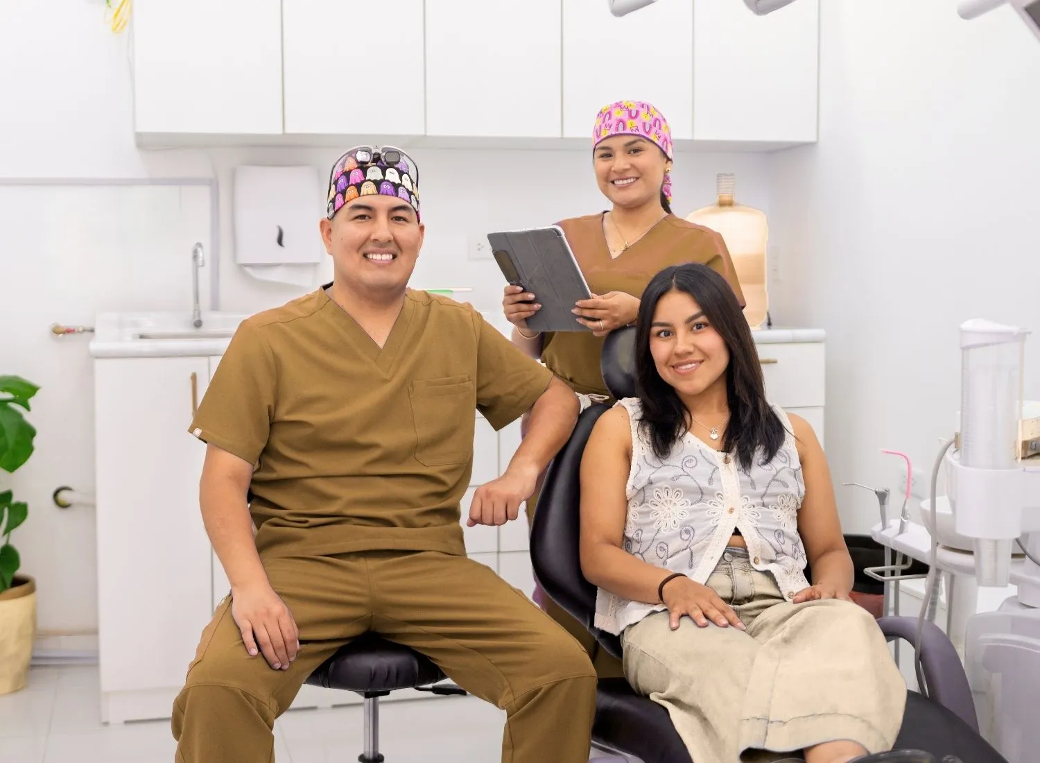 Female patient sits in dental chair while two dentists in brown scrubs smile at camera in clinic.