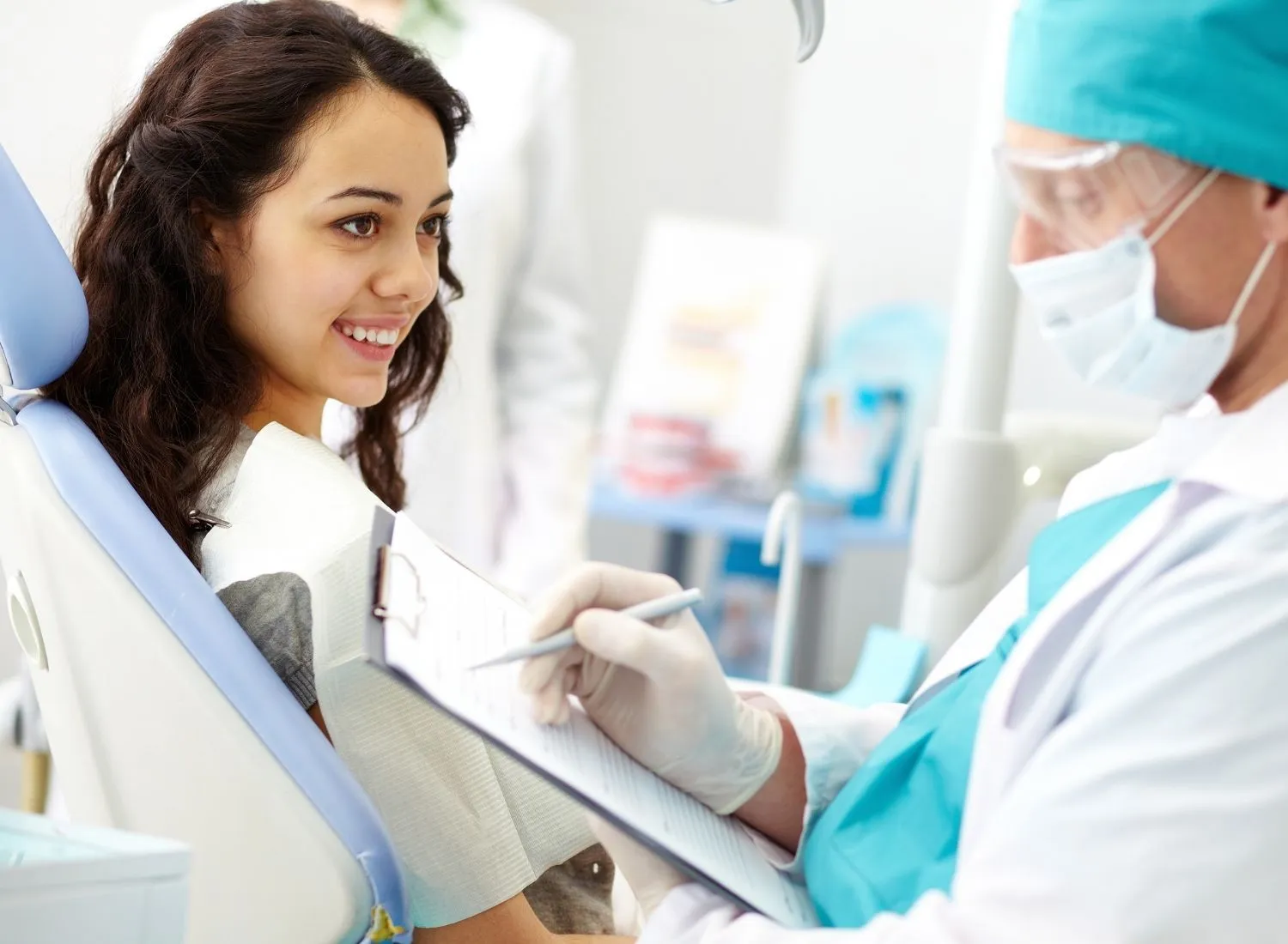 Smiling young woman in dental chair talking to masked dentist writing on clipboard.