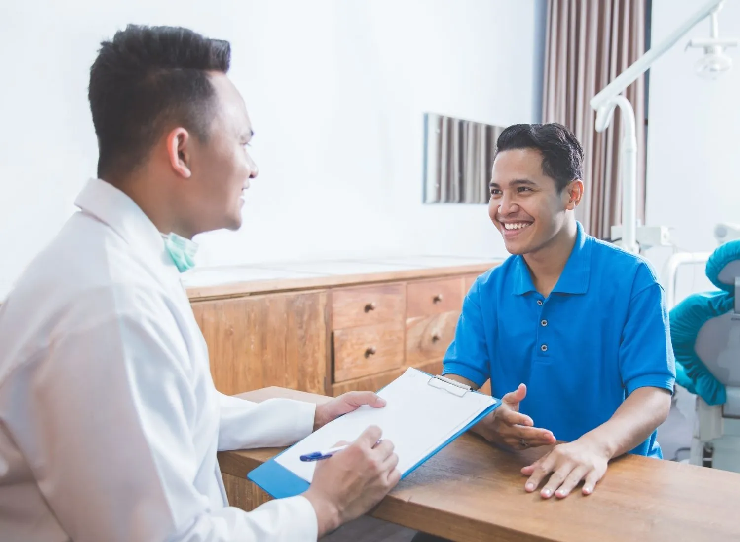 Dentist in white coat holding clipboard talking to smiling male patient in blue polo shirt.