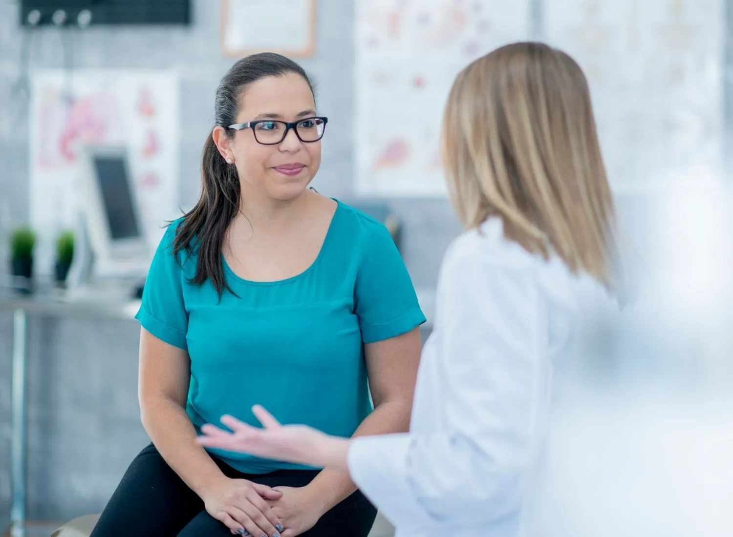 Woman in glasses and teal shirt listening to a healthcare professional in a white coat.