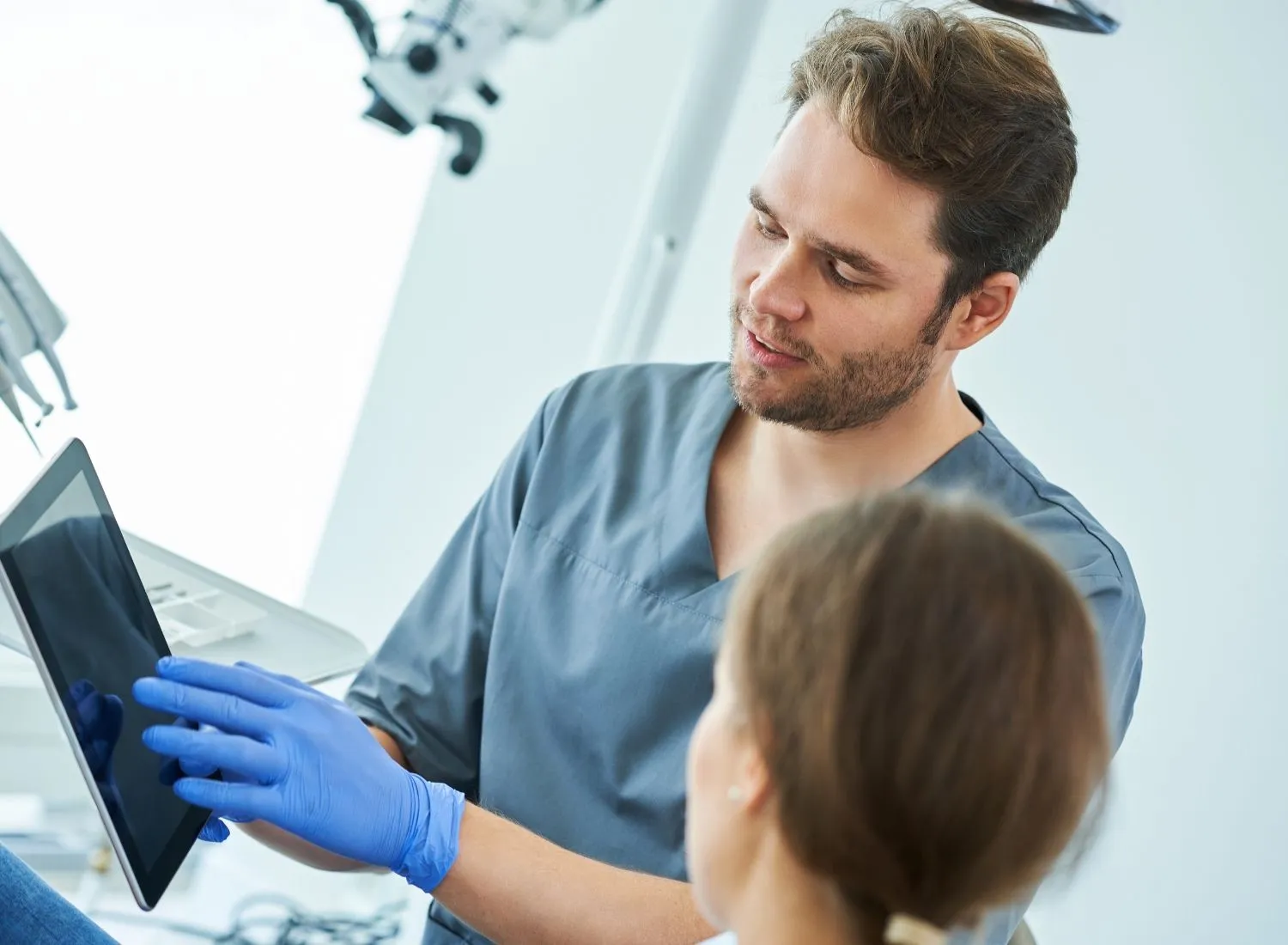 Dentist with gloves showing a tablet screen to a female patient in a dental clinic.