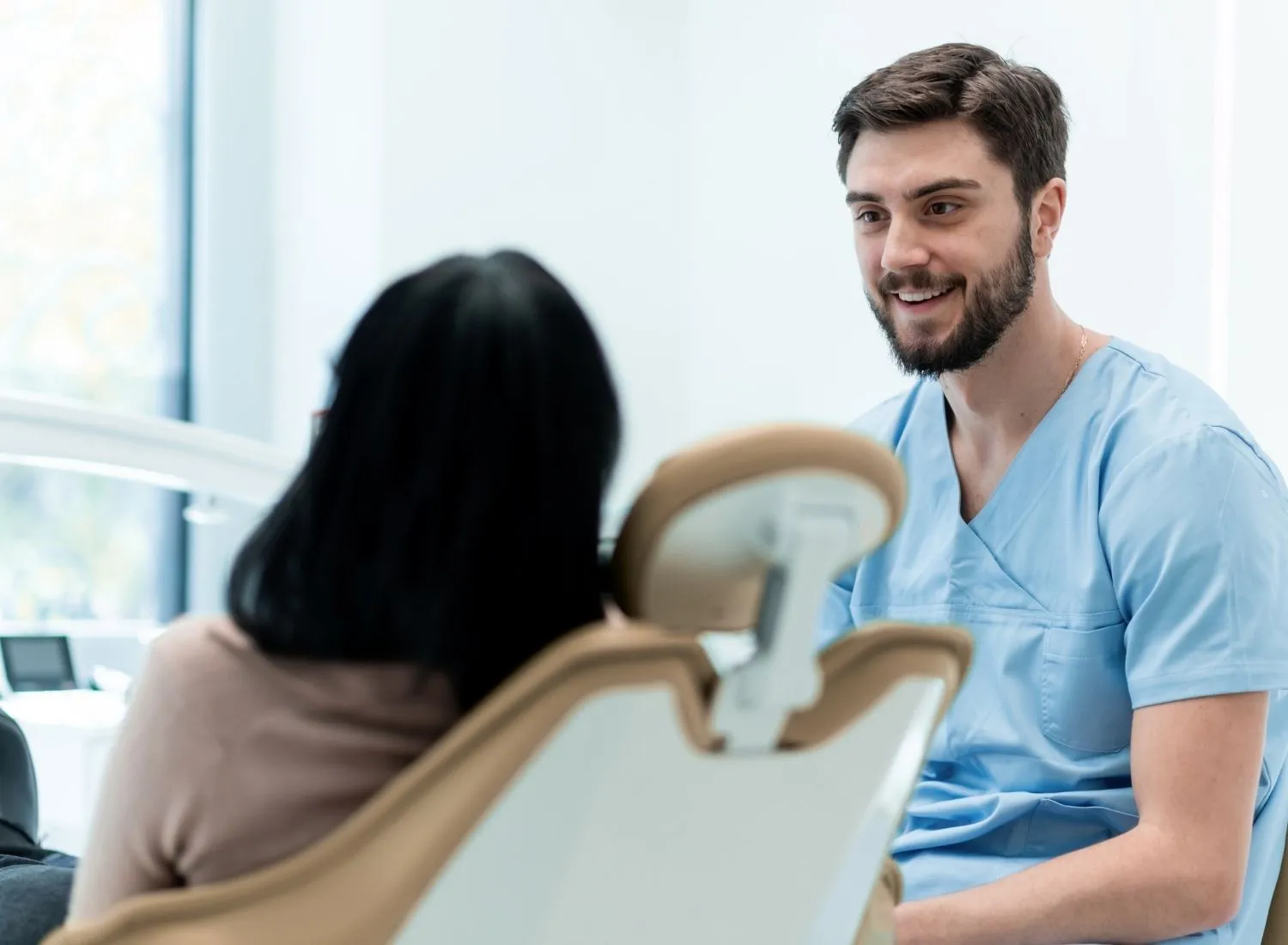 Male dentist in blue scrubs smiling and talking to a patient seated in a dental chair.