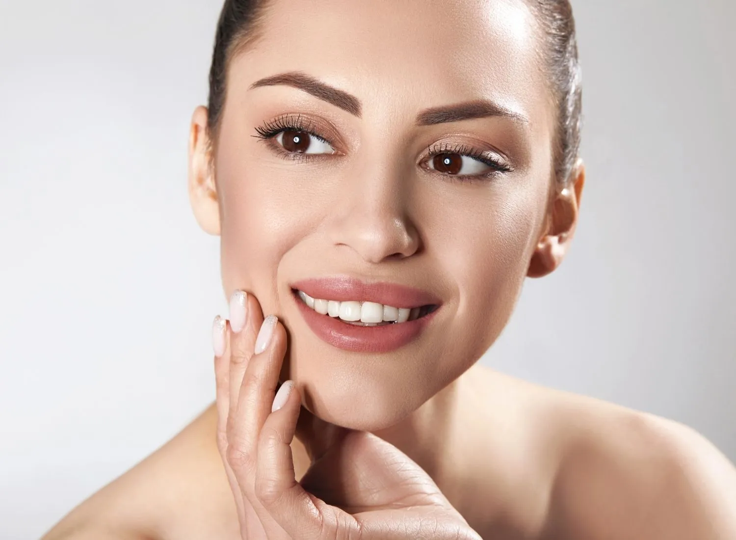 Close-up of a smiling woman with clear skin touching her cheek against a light background.
