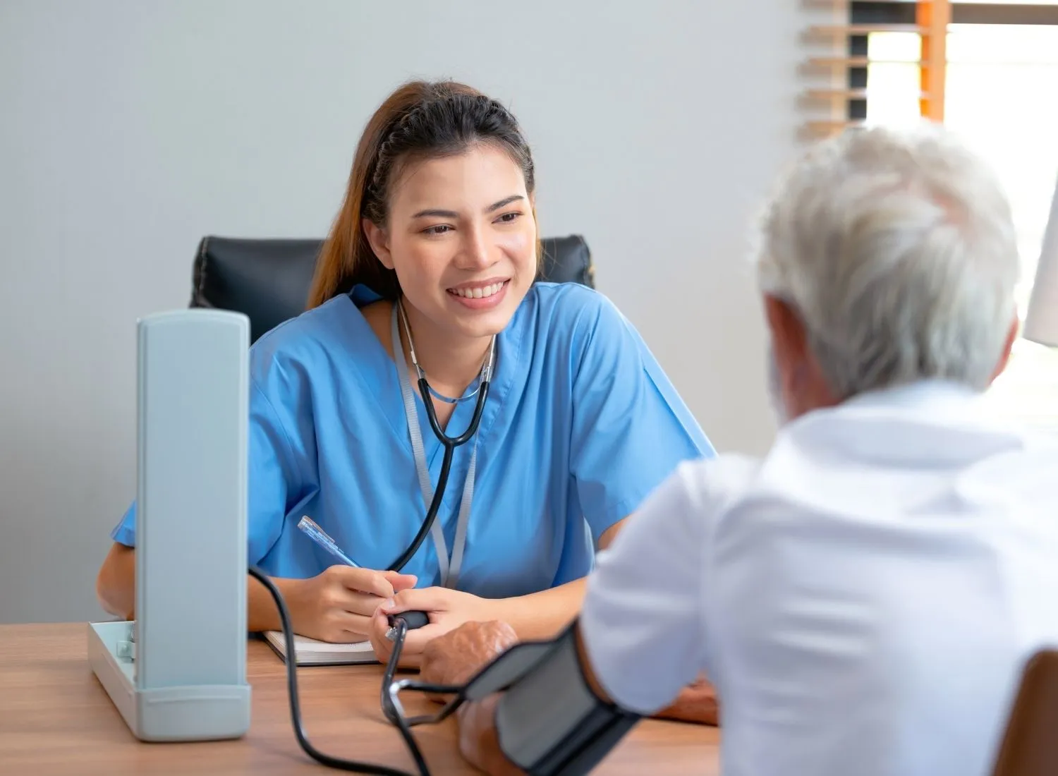 Nurse in blue scrubs smiling while taking blood pressure of elderly man in white shirt.