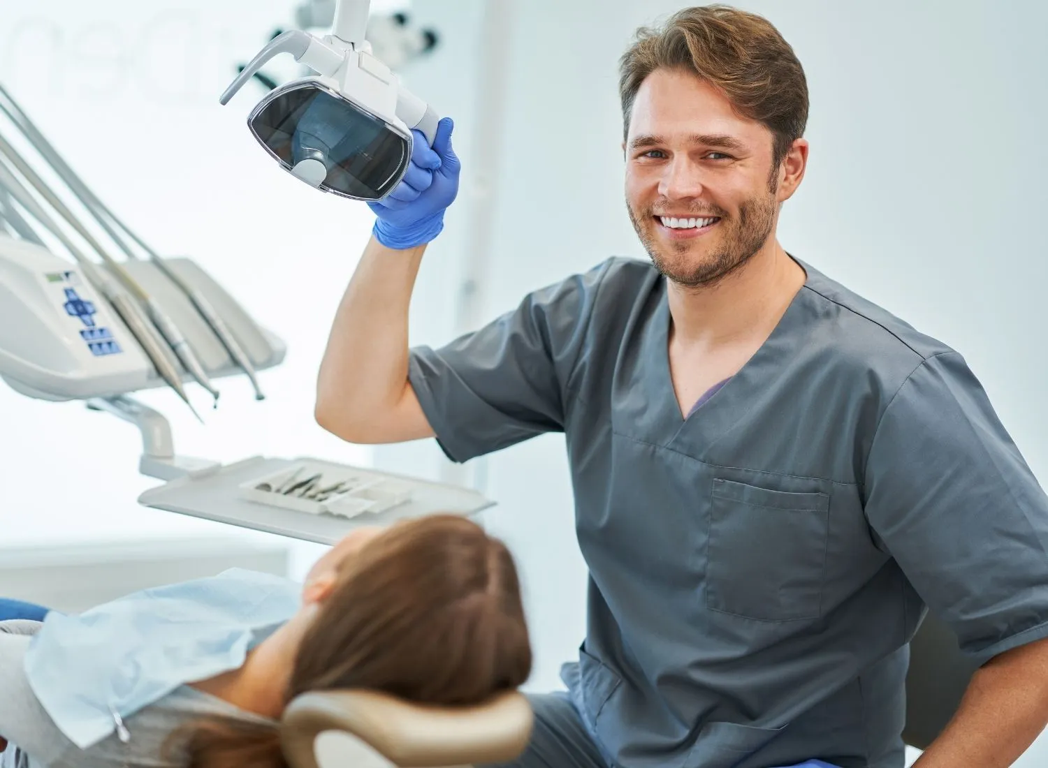Smiling male dentist in gray scrubs holding a dental light above a patient in the dentist chair.