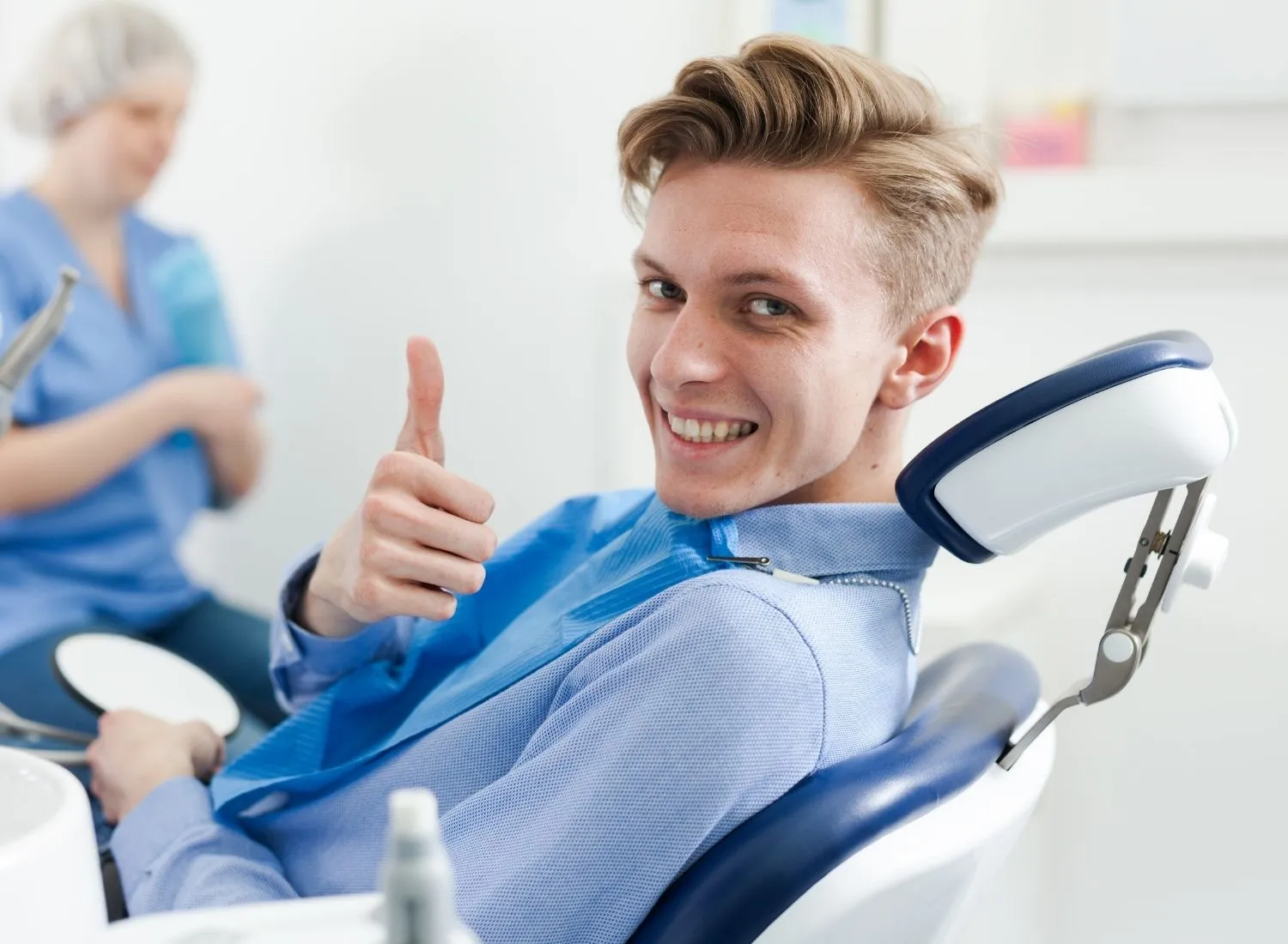 Young man in dental chair giving thumbs up and smiling with dental assistant in background.