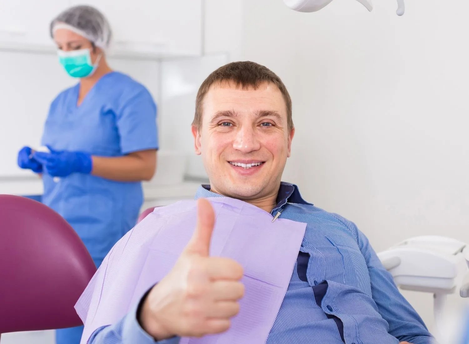 Smiling man in dental chair giving thumbs up with a dental assistant in blue scrubs and mask in background.